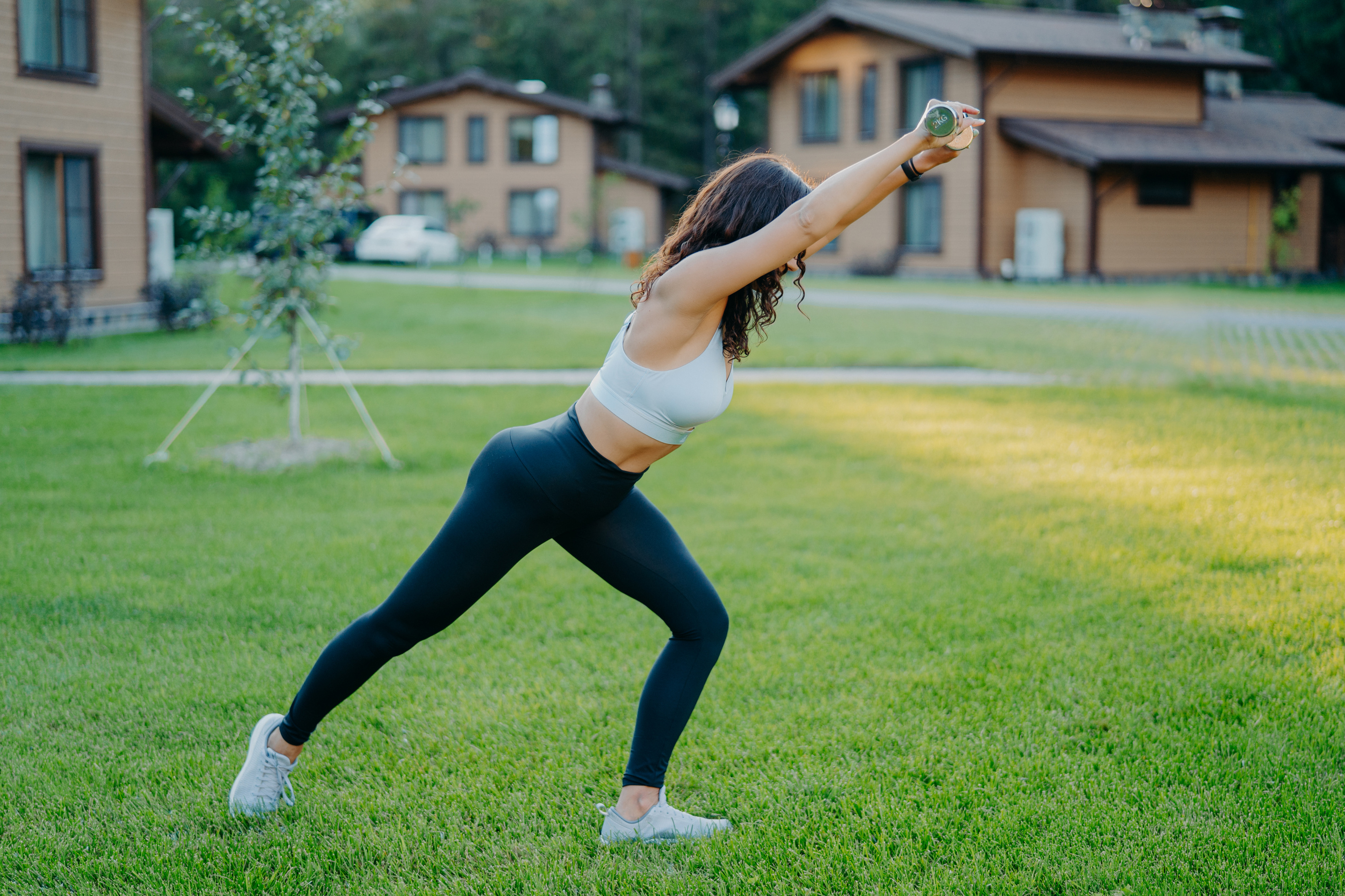 Horizontal Shot of Active Slim Woman Does Stretching Exercises with Dumbbells, Dressed in Cropped Top and Leggings, Enjoys Morning Summer Workout on Green Grass. Bodybuilding, Flexibility Concept